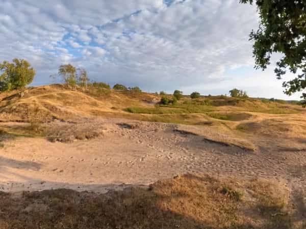 Zandpad langs droge heuvels en bomen onder een wolkenlucht